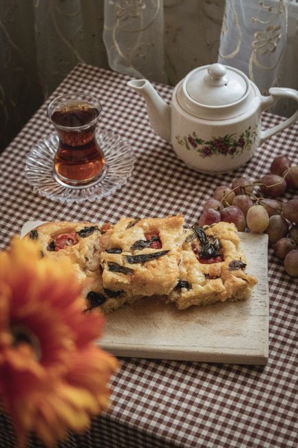 A close-up scene of a table set for a casual home snack or tea break, featuring a checkered brown and white tablecloth. On the table, there is a white ceramic teapot decorated with a floral pattern, a glass of dark tea placed on a glass saucer, and a wooden cutting board with several pieces of homemade fruit-flavored cake or pastry topped with berries. To the right, a small bunch of red and green grapes rests on the table. In the foreground, a blurred orange flower is visible, adding a splash of color. The background shows a light, patterned curtain, suggesting a cozy kitchen or dining area. The focus is on the food and drink accessories, creating a warm, inviting atmosphere often associated with home relaxation or casual gatherings, relevant to the context of household activities such as packing or moving preparations.