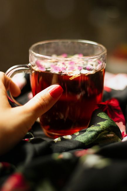 A close-up of a person's hand with neatly manicured nails holding a clear glass mug filled with a dark red herbal tea, topped with pink and white flower petals. The mug is held above a soft fabric surface with a floral pattern in dark colors, possibly part of a blanket or clothing. The scene appears to be indoors with warm lighting. This image is related to house moving services provided by Earls Court Man and Van, emphasizing the comfort and relaxation during a home relocation, such as taking a break with a hot beverage amidst packing and furniture transport activities.
