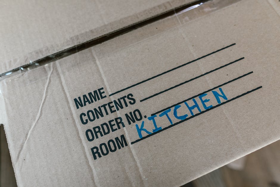 Close-up of a cardboard moving box with printed labels indicating sections for name, contents, and order number, with 'Kitchen' written in blue marker on the 'Room' line. The box is sealed with clear packing tape and appears to be packed with household items for a home relocation. The background shows other boxes and packing materials, suggesting an inside property environment during the packing or loading process, typical of furniture transport and packing and moving activities. The image naturally aligns with the services provided by Earls Court Man and Van in house removals, specifically during cabinet packing and loading procedures for a house or flat move on Gloucester Road.