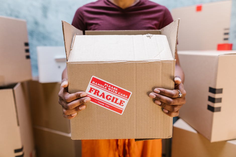 A person holding an open cardboard box marked with a red and white 'Fragile' handling sticker, surrounded by other packed boxes in a warehouse or moving storage area. The individual’s hands, with dark skin and a silver ring on the left ring finger, grip the sides of the box. In the background, additional closed cardboard boxes are stacked on a surface, some with black tape sealing them, indicating preparations for a home relocation. The lighting is bright and even, highlighting the textures of the cardboard and packing materials. The setting suggests an indoor environment where packing and moving logistics are underway, consistent with furniture transport and packing procedures associated with house removals, serviced by companies like Earls Court Man and Van.