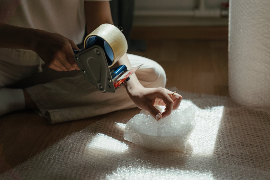 A person sitting on the floor inside a home, using a handheld tape dispenser to apply packing tape from a roll onto a piece of crumpled bubble wrap. The individual is dressed in light clothing and is surrounded by packing materials such as cardboard boxes and additional bubble wrap. The scene shows the early stage of a packing and moving process, with the person's hands visible, focusing on securely sealing items for transportation. The setting includes a wooden floor and natural light illuminating the workspace, with the surrounding environment indicating a residential interior prepared for a house relocation. This detailed image reflects careful packing activities typically associated with professional removals, as offered by Earls Court Man and Van.