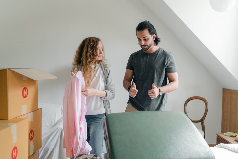 Inside a residential interior, a man and a woman are engaged in a packing and moving process. The woman, with curly red hair, is holding a pink and white striped bedding or fabric, while the man, with dark hair tied back, is gesturing with both thumbs up. The room contains several packed cardboard boxes, some stacked near a white sofa, indicating an ongoing home relocation. The surrounding environment includes a wooden chair with a rounded back adhered to the wall, and a sloped ceiling with a window allowing natural light to fill the space. Visible equipment such as a trolley or cart may be present, supporting furniture transport and loading activities. This scene exemplifies a careful packing and moving effort, supported by professional services like those from Earls Court Man and Van, focused on efficient furniture transport and relocation logistics within a domestic setting.