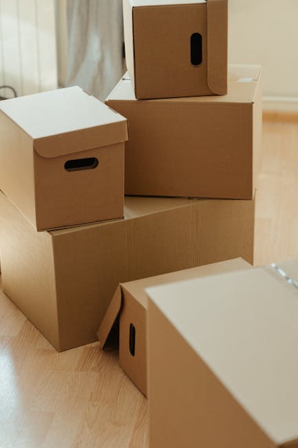 A person sitting on the floor inside a home, using a handheld tape dispenser to apply packing tape from a roll onto a piece of crumpled bubble wrap. The individual is dressed in light clothing and is surrounded by packing materials such as cardboard boxes and additional bubble wrap. The scene shows the early stage of a packing and moving process, with the person's hands visible, focusing on securely sealing items for transportation. The setting includes a wooden floor and natural light illuminating the workspace, with the surrounding environment indicating a residential interior prepared for a house relocation. This detailed image reflects careful packing activities typically associated with professional removals, as offered by Earls Court Man and Van.