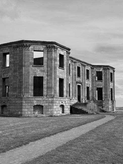 A black-and-white image of an old, vacant building with classical architectural elements, including tall, fluted columns flanking the front entrance and large rectangular windows without glass or frames. The structure appears to be in a state of disrepair, with visible weathering on the stone facade and some structural deterioration. The building is situated on a grassy area with a narrow, curved pathway leading up to the steps at the entrance. In the background, the sky is cloudy, and there are no other structures visible nearby. The scene is outdoors, and the overall setting suggests a historic or abandoned site that could be of interest in discussions about building restoration or secure storage during home relocations. The label references a removals context based on service relevance, though visually it depicts an uninhabited, classical-style building.