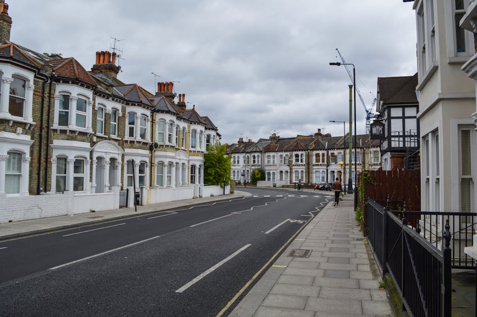 Street view of a residential area near Earls Court Road featuring a row of Victorian-style terraced houses with white facades, bay windows, red-tiled roofs, and chimneys under an overcast sky. The pavement runs alongside the buildings, with a black metal fence and potted plants visible on the right. A person is walking along the sidewalk, and a crane and construction site can be seen further down the street. The scene is calm and typical of urban home relocation surroundings, with no moving activity visible but conveying the context of local house removals and furniture transport possibly organized by Earls Court Man and Van, as suggested by the page title.
